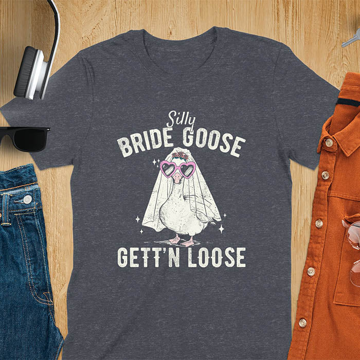 A t-shirt with a goose dressed as a bride with heart sunglasses and a veil. Text: "Silly BRIDE GOOSE" above and "GETT'N LOOSE" below. This funny bride shirt features a silly goose for getting ready bridal. A bride crewneck and engaged gift, a fiancé gift for honeymoon outfit or honeymoon gifts. A married couple shirt or new bride shirt, a fun engagement announce idea. A couple's tee-shirt and funny goose gift.