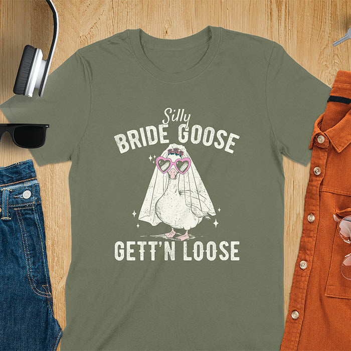 A t-shirt with a goose dressed as a bride with heart sunglasses and a veil. Text: "Silly BRIDE GOOSE" above and "GETT'N LOOSE" below. This funny bride shirt features a silly goose for getting ready bridal. A bride crewneck and engaged gift, a fiancé gift for honeymoon outfit or honeymoon gifts. A married couple shirt or new bride shirt, a fun engagement announce idea. A couple's tee-shirt and funny goose gift.