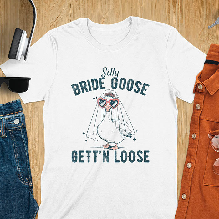 A t-shirt with a goose dressed as a bride with heart sunglasses and a veil. Text: "Silly BRIDE GOOSE" above and "GETT'N LOOSE" below. This funny bride shirt features a silly goose for getting ready bridal. A bride crewneck and engaged gift, a fiancé gift for honeymoon outfit or honeymoon gifts. A married couple shirt or new bride shirt, a fun engagement announce idea. A couple's tee-shirt and funny goose gift.