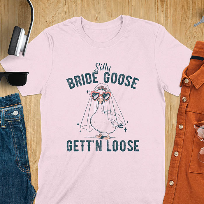 A t-shirt with a goose dressed as a bride with heart sunglasses and a veil. Text: "Silly BRIDE GOOSE" above and "GETT'N LOOSE" below. This funny bride shirt features a silly goose for getting ready bridal. A bride crewneck and engaged gift, a fiancé gift for honeymoon outfit or honeymoon gifts. A married couple shirt or new bride shirt, a fun engagement announce idea. A couple's tee-shirt and funny goose gift.