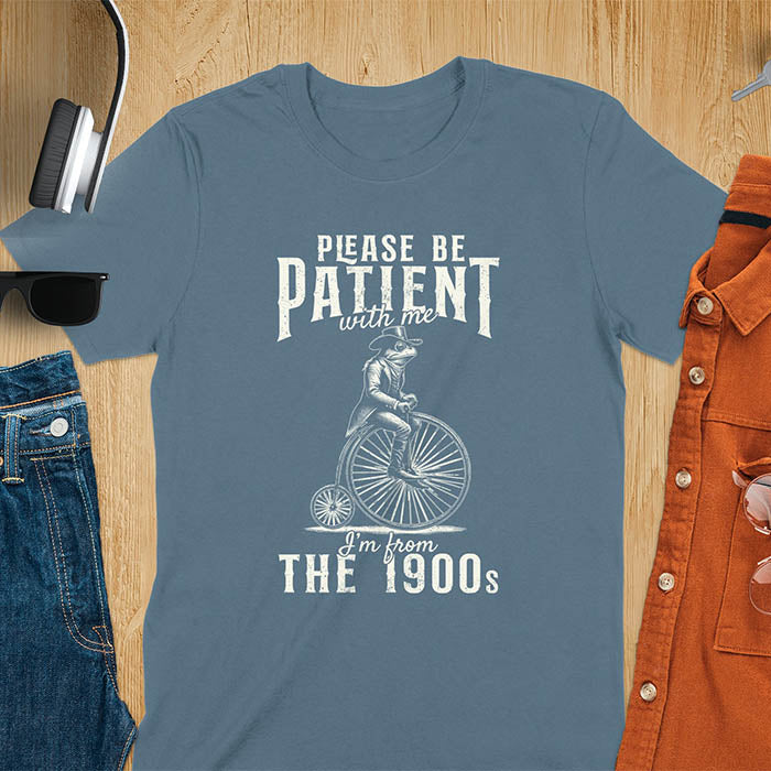 A t-shirt with a frog riding a penny-farthing bicycle. Text: "PLEASE BE PATIENT with me" above and "I'm from THE 1900s" below. This The 1900s shirt has a vintage graphic tee and retro aesthetic. A genz Meme shirt and weird silly shirt, a funny gift friend. A gift for husband, funny grandpa gift, or dad tee-shirt. An animal person gift, a 90s country shirt, a mom gift option.