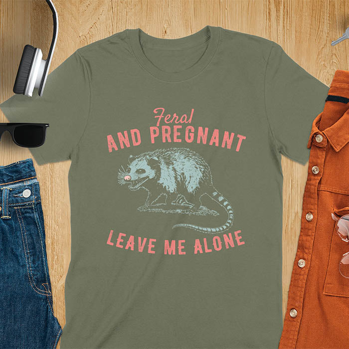 An upset-looking opossum showing its fangs. Text: "Feral AND PREGNANT LEAVE ME ALONE". This funny maternity tee for pregnancy humor is ideal for expecting mom gift & mommy to be gift. Supports feral pregnancy, pregnancy announce, expecting baby, pregnancy keepsake. A unique new mama shirt, maternity apparel & gift mom hers.