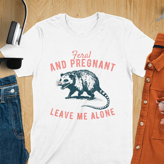 An upset-looking opossum showing its fangs. Text: "Feral AND PREGNANT LEAVE ME ALONE". This funny maternity tee for pregnancy humor is ideal for expecting mom gift & mommy to be gift. Supports feral pregnancy, pregnancy announce, expecting baby, pregnancy keepsake. A unique new mama shirt, maternity apparel & gift mom hers.