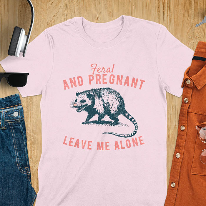 An upset-looking opossum showing its fangs. Text: "Feral AND PREGNANT LEAVE ME ALONE". This funny maternity tee for pregnancy humor is ideal for expecting mom gift & mommy to be gift. Supports feral pregnancy, pregnancy announce, expecting baby, pregnancy keepsake. A unique new mama shirt, maternity apparel & gift mom hers.