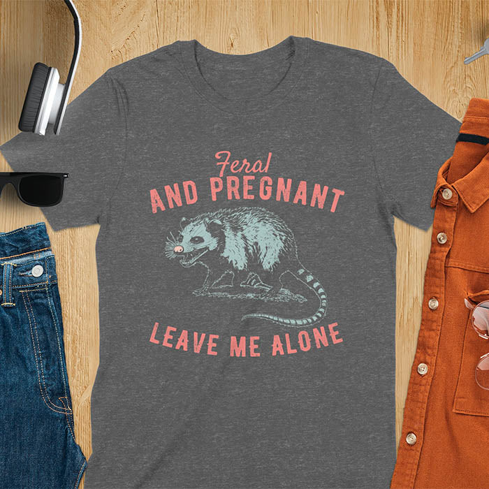 An upset-looking opossum showing its fangs. Text: "Feral AND PREGNANT LEAVE ME ALONE". This funny maternity tee for pregnancy humor is ideal for expecting mom gift & mommy to be gift. Supports feral pregnancy, pregnancy announce, expecting baby, pregnancy keepsake. A unique new mama shirt, maternity apparel & gift mom hers.
