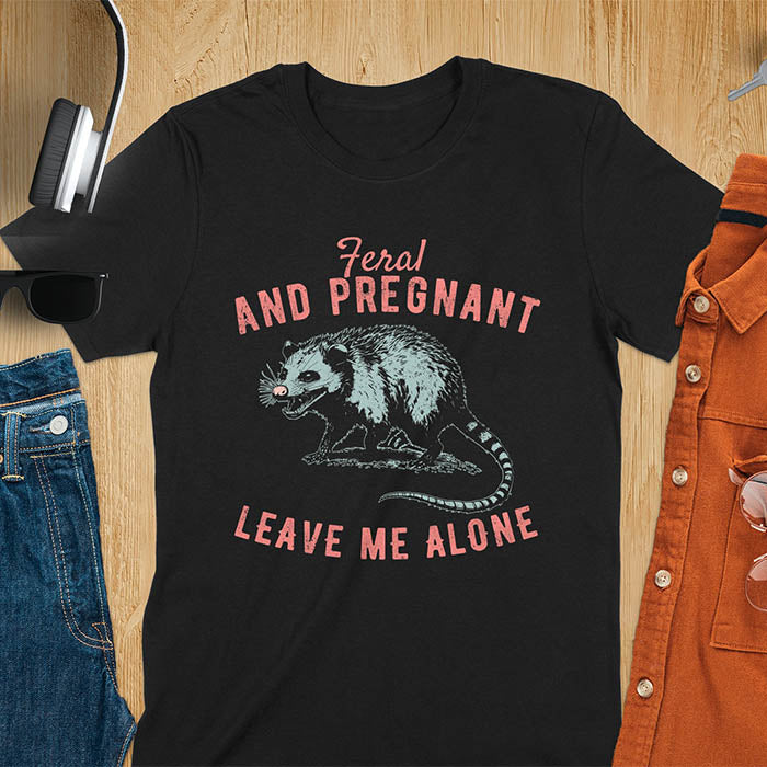 An upset-looking opossum showing its fangs. Text: "Feral AND PREGNANT LEAVE ME ALONE". This funny maternity tee for pregnancy humor is ideal for expecting mom gift & mommy to be gift. Supports feral pregnancy, pregnancy announce, expecting baby, pregnancy keepsake. A unique new mama shirt, maternity apparel & gift mom hers.
