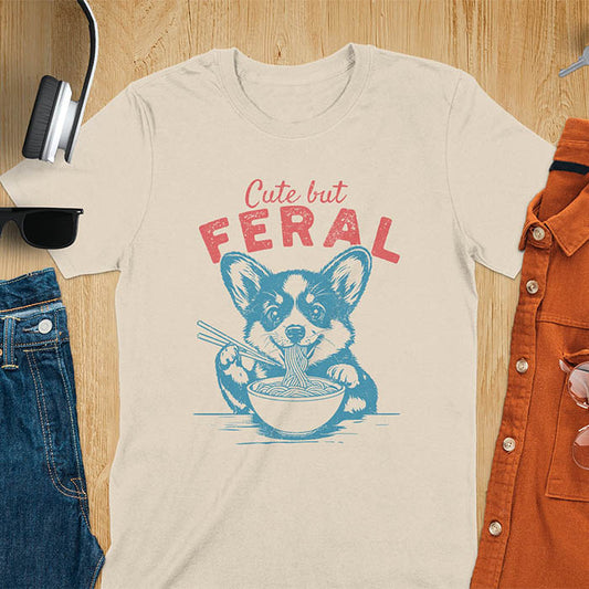 A t-shirt with a corgi eating noodles with chopsticks. Text: Cute but FERAL above. This funny dog shirt features kawaiiness cuteness, a meme graphic tee. A retro dog shirt and vintage dog tee, an animal person gift or gift mom hers. A funny unhinge shirt or weird funny shirt for a dog dad shirt. A comfortable t-shirts option with a trendy tshirt design, a funny animal meme.