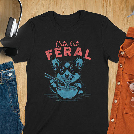 A t-shirt with a corgi eating noodles with chopsticks. Text: Cute but FERAL above. This funny dog shirt features kawaiiness cuteness, a meme graphic tee. A retro dog shirt and vintage dog tee, an animal person gift or gift mom hers. A funny unhinge shirt or weird funny shirt for a dog dad shirt. A comfortable t-shirts option with a trendy tshirt design, a funny animal meme.