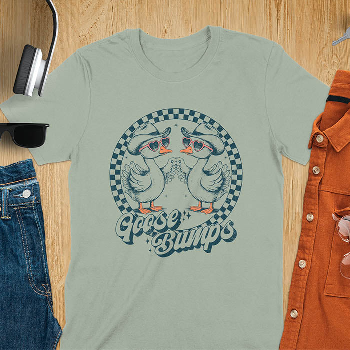 A t-shirt with two geese in cowboy hats and heart sunglasses touching hands, inside a checkered circle. Text "Goose Bumps" is below them on a curved banner. A silly goose crewneck, sarcastic, Funny Sarcastic Meme Tee-shirt, Retro Western Goose Shirt, Snarky Sassy Saying Tshirt, unhinged tee-shirt, weirdcore meme. Ideal gift for goose lover, cowboy cowgirl gift, animal person, gift mom hers. Features kawaiiness cuteness, cute animal clothe, Vintage Silly Humorous Tee Gift.