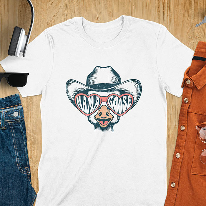 A t-shirt featuring a goose wearing a cowboy hat and oversized pink heart-shaped sunglasses. The pink lenses of the sunglasses display the words Mama Goose in a stylized font. The goose graphic is centered on the shirt and has a vintage distressed texture.