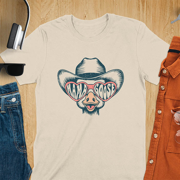 A t-shirt featuring a goose wearing a cowboy hat and oversized pink heart-shaped sunglasses. The pink lenses of the sunglasses display the words Mama Goose in a stylized font. The goose graphic is centered on the shirt and has a vintage distressed texture.