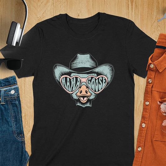 A t-shirt featuring a goose wearing a cowboy hat and oversized pink heart-shaped sunglasses. The pink lenses of the sunglasses display the words Mama Goose in a stylized font. The goose graphic is centered on the shirt and has a vintage distressed texture.