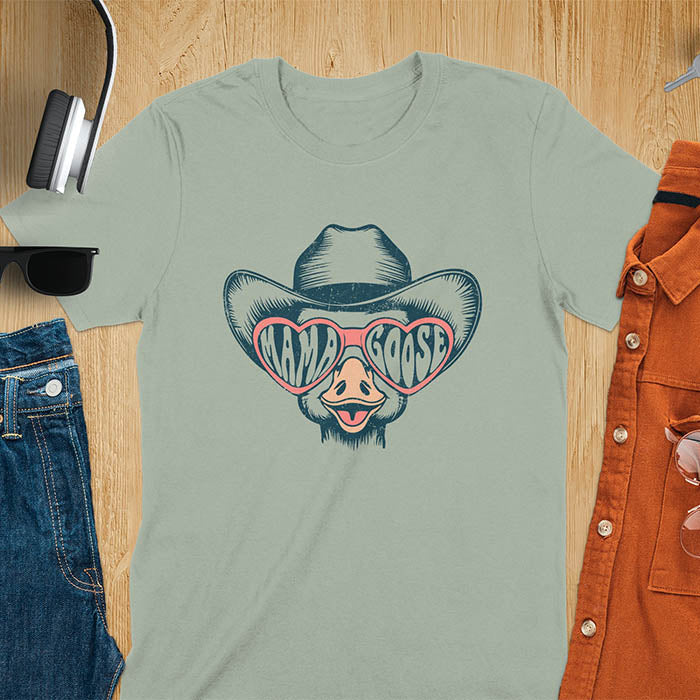 A t-shirt featuring a goose wearing a cowboy hat and oversized pink heart-shaped sunglasses. The pink lenses of the sunglasses display the words Mama Goose in a stylized font. The goose graphic is centered on the shirt and has a vintage distressed texture.