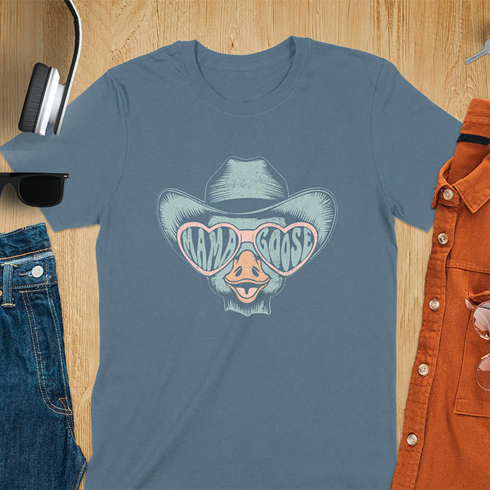 A t-shirt featuring a goose wearing a cowboy hat and oversized pink heart-shaped sunglasses. The pink lenses of the sunglasses display the words Mama Goose in a stylized font. The goose graphic is centered on the shirt and has a vintage distressed texture.