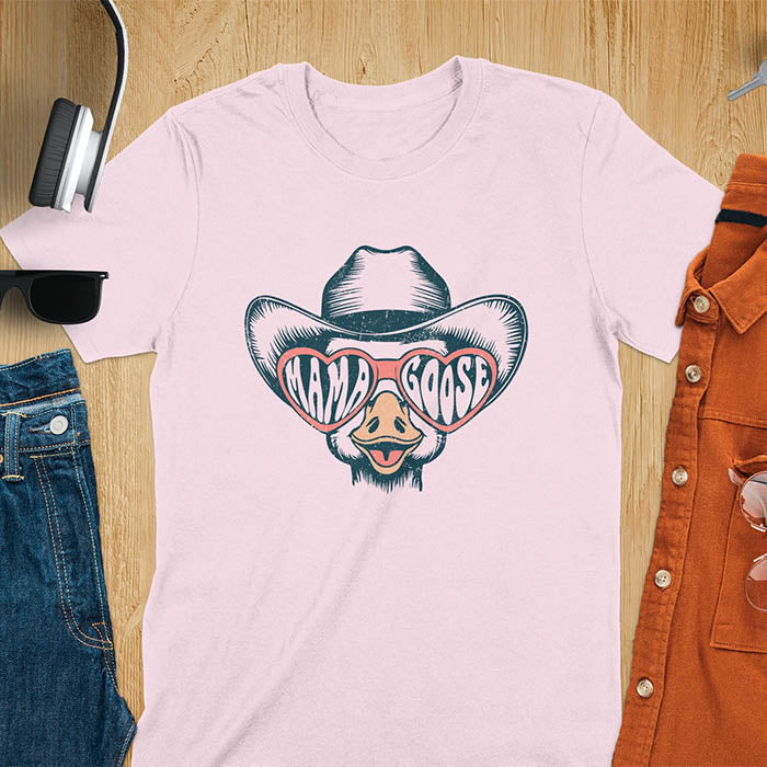 A t-shirt featuring a goose wearing a cowboy hat and oversized pink heart-shaped sunglasses. The pink lenses of the sunglasses display the words Mama Goose in a stylized font. The goose graphic is centered on the shirt and has a vintage distressed texture.