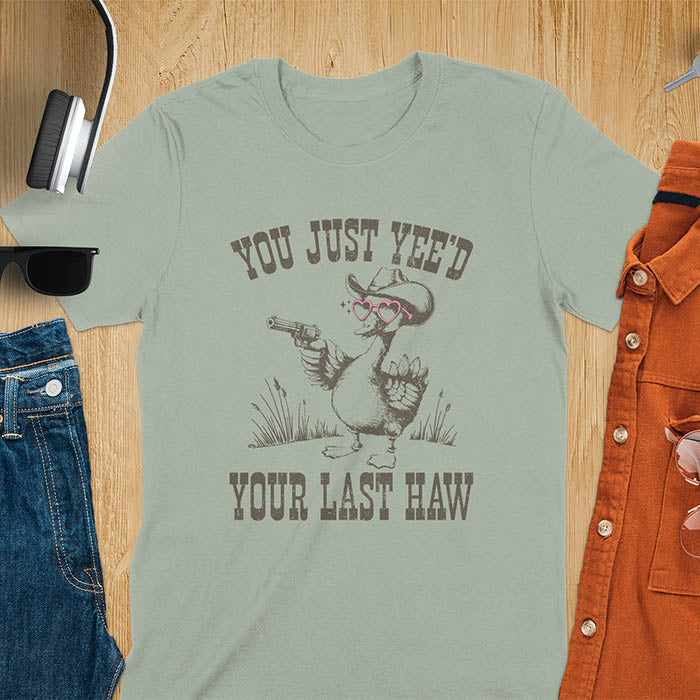 A t-shirt with a goose wearing a cowboy hat and sunglasses, holding a pistol. Text: "YOU JUST YEE'D" above and "YOUR LAST HAW" below. This funny western shirt features a silly goose shirt or goose lovers gift. A cowboy tshirt or cowgirl shirt, a western style tee and western shirt. A retro graphic tee and weirdcore tshirt, an animal person gift or animal lover gag. A mom gift alternative.