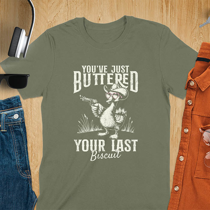 A t-shirt with a goose wearing a cowboy hat and sunglasses, holding a pistol. Text: "YOU'VE JUST BUTTERED" above and "YOUR LAST Biscuit" below. This funny cowboy shirt features a silly goose shirt or goose lovers gift. A cowgirl t-shirt and country girl shirt, a western girl shirt. A retro graphic tee and weirdcore tee-shirt, an animal person gift or animal lover gag. A mom gift alternative, a last biscuit shirt with a sarcastic quote.