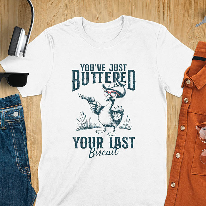 A t-shirt with a goose wearing a cowboy hat and sunglasses, holding a pistol. Text: "YOU'VE JUST BUTTERED" above and "YOUR LAST Biscuit" below. This funny cowboy shirt features a silly goose shirt or goose lovers gift. A cowgirl t-shirt and country girl shirt, a western girl shirt. A retro graphic tee and weirdcore tee-shirt, an animal person gift or animal lover gag. A mom gift alternative, a last biscuit shirt with a sarcastic quote.