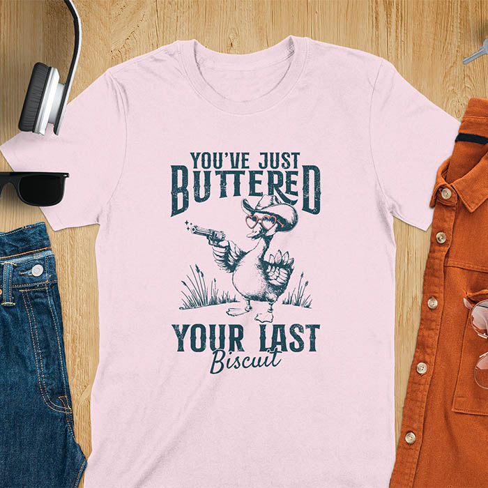 A t-shirt with a goose wearing a cowboy hat and sunglasses, holding a pistol. Text: "YOU'VE JUST BUTTERED" above and "YOUR LAST Biscuit" below. This funny cowboy shirt features a silly goose shirt or goose lovers gift. A cowgirl t-shirt and country girl shirt, a western girl shirt. A retro graphic tee and weirdcore tee-shirt, an animal person gift or animal lover gag. A mom gift alternative, a last biscuit shirt with a sarcastic quote.