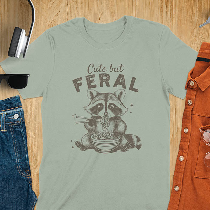 A t-shirt with a raccoon eating ramen with chopsticks. Text above: "Cute but". Text below: "FERAL". This Cute But Feral Shirt is a Funny Raccoon T-shirt, Vintage Retro Graphic Ramen Tee-shirt. A Silly Meme Tee, Hilarious Gag Gift, funny unhinge shirt, sarcastic raccoon, trendy tshirt design. Features kawaiiness cuteness. Ideal for animal person gift, raccoon lover shirt, gift mom hers, feral mom t shirt. A vintage inspired Animal Lover Tshirt, almost feral shirt.
