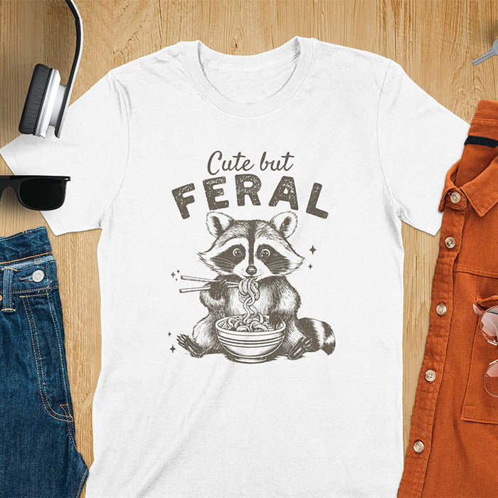 A t-shirt with a raccoon eating ramen with chopsticks. Text above: "Cute but". Text below: "FERAL". This Cute But Feral Shirt is a Funny Raccoon T-shirt, Vintage Retro Graphic Ramen Tee-shirt. A Silly Meme Tee, Hilarious Gag Gift, funny unhinge shirt, sarcastic raccoon, trendy tshirt design. Features kawaiiness cuteness. Ideal for animal person gift, raccoon lover shirt, gift mom hers, feral mom t shirt. A vintage inspired Animal Lover Tshirt, almost feral shirt.