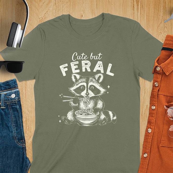 A t-shirt with a raccoon eating ramen with chopsticks. Text above: "Cute but". Text below: "FERAL". This Cute But Feral Shirt is a Funny Raccoon T-shirt, Vintage Retro Graphic Ramen Tee-shirt. A Silly Meme Tee, Hilarious Gag Gift, funny unhinge shirt, sarcastic raccoon, trendy tshirt design. Features kawaiiness cuteness. Ideal for animal person gift, raccoon lover shirt, gift mom hers, feral mom t shirt. A vintage inspired Animal Lover Tshirt, almost feral shirt.