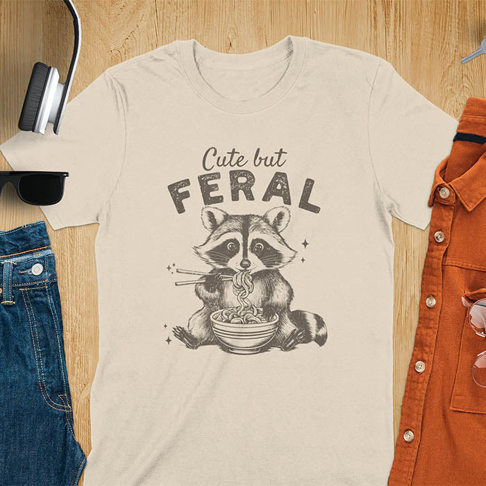 A t-shirt with a raccoon eating ramen with chopsticks. Text above: "Cute but". Text below: "FERAL". This Cute But Feral Shirt is a Funny Raccoon T-shirt, Vintage Retro Graphic Ramen Tee-shirt. A Silly Meme Tee, Hilarious Gag Gift, funny unhinge shirt, sarcastic raccoon, trendy tshirt design. Features kawaiiness cuteness. Ideal for animal person gift, raccoon lover shirt, gift mom hers, feral mom t shirt. A vintage inspired Animal Lover Tshirt, almost feral shirt.