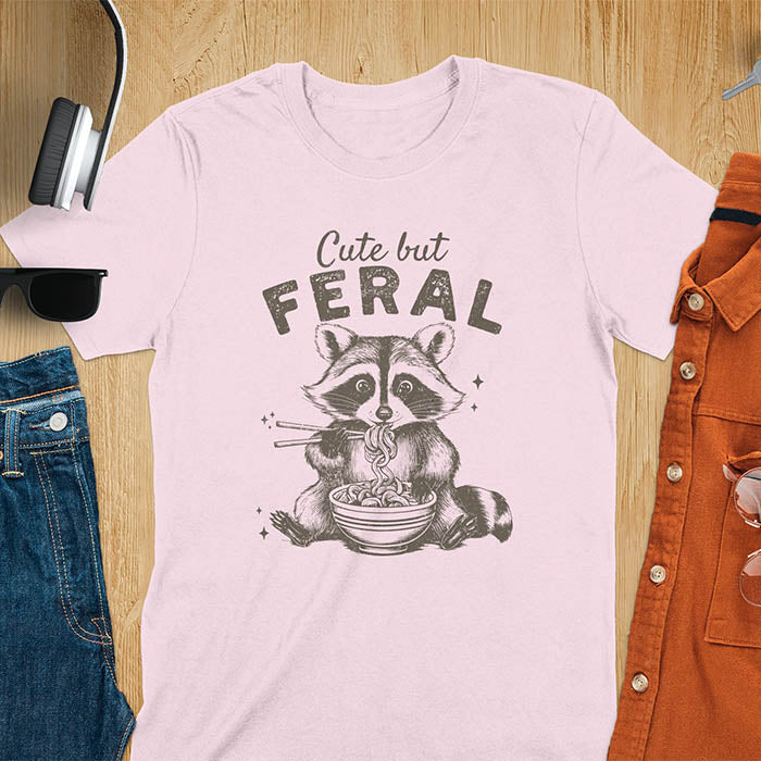 A t-shirt with a raccoon eating ramen with chopsticks. Text above: "Cute but". Text below: "FERAL". This Cute But Feral Shirt is a Funny Raccoon T-shirt, Vintage Retro Graphic Ramen Tee-shirt. A Silly Meme Tee, Hilarious Gag Gift, funny unhinge shirt, sarcastic raccoon, trendy tshirt design. Features kawaiiness cuteness. Ideal for animal person gift, raccoon lover shirt, gift mom hers, feral mom t shirt. A vintage inspired Animal Lover Tshirt, almost feral shirt.