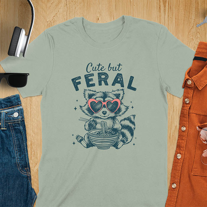 A t-shirt with a raccoon in heart sunglasses eating ramen. Text: "Cute but FERAL". A Funny Cute But Feral Raccoon Shirt, Vintage Retro Graphic Ramen Tee, Silly Sarcastic Tshirt, Hilarious Gag Gift. Unhinged, trash panda, trendy design. Features kawaiiness cuteness. Great gift for animal person, raccoon lover, mom, feral mom. Vintage inspired Animal Lover T-shirt, almost feral shirt.