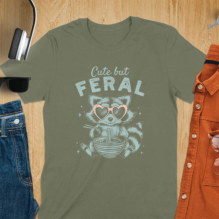 A t-shirt with a raccoon in heart sunglasses eating ramen. Text: "Cute but FERAL". A Funny Cute But Feral Raccoon Shirt, Vintage Retro Graphic Ramen Tee, Silly Sarcastic Tshirt, Hilarious Gag Gift. Unhinged, trash panda, trendy design. Features kawaiiness cuteness. Great gift for animal person, raccoon lover, mom, feral mom. Vintage inspired Animal Lover T-shirt, almost feral shirt.