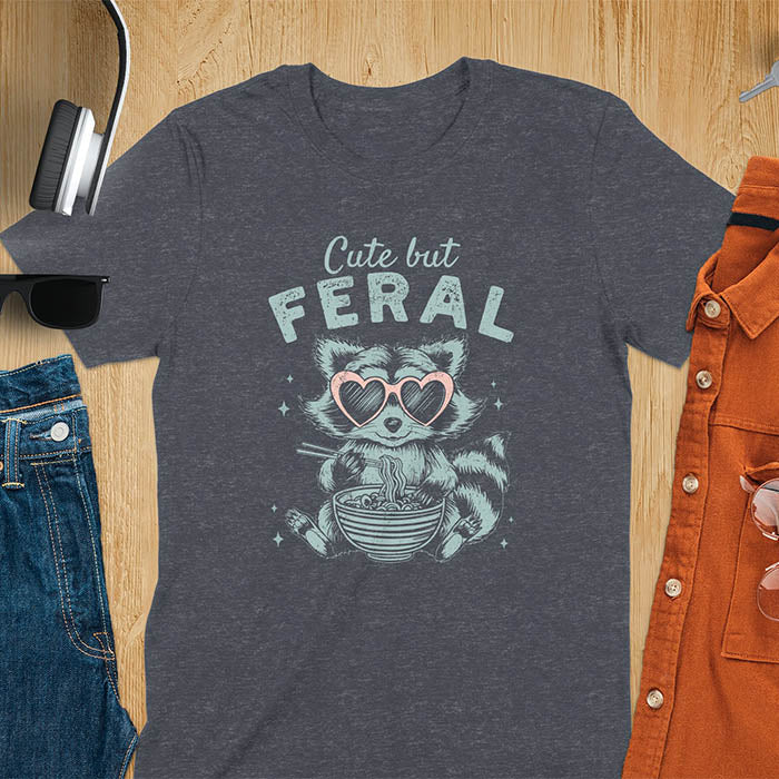 A t-shirt with a raccoon in heart sunglasses eating ramen. Text: "Cute but FERAL". A Funny Cute But Feral Raccoon Shirt, Vintage Retro Graphic Ramen Tee, Silly Sarcastic Tshirt, Hilarious Gag Gift. Unhinged, trash panda, trendy design. Features kawaiiness cuteness. Great gift for animal person, raccoon lover, mom, feral mom. Vintage inspired Animal Lover T-shirt, almost feral shirt.