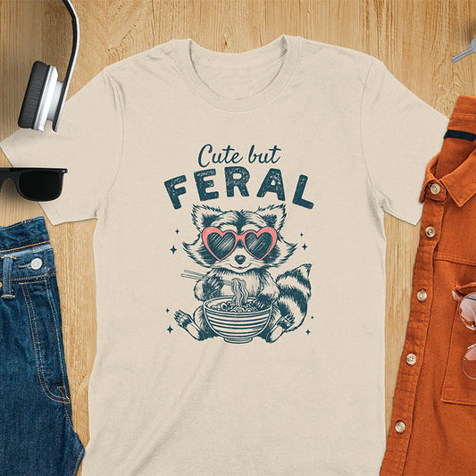 A t-shirt with a raccoon in heart sunglasses eating ramen. Text: "Cute but FERAL". A Funny Cute But Feral Raccoon Shirt, Vintage Retro Graphic Ramen Tee, Silly Sarcastic Tshirt, Hilarious Gag Gift. Unhinged, trash panda, trendy design. Features kawaiiness cuteness. Great gift for animal person, raccoon lover, mom, feral mom. Vintage inspired Animal Lover T-shirt, almost feral shirt.