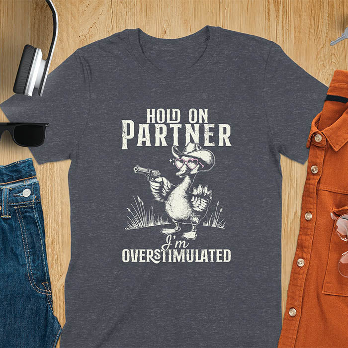 A t-shirt with a goose wearing a cowboy hat and holding a pistol. Text: "HOLD ON PARTNER" above and "I'm OVERSTIMULATED" below. This cowboy frog shirt is a funny meme shirt and oddly specific t shirt. A retro graphic tee and unhinged tshirt, a funny animal shirt. An animal person gift or goose lovers gift, an animal lover gag or mom gift for overstimulated memes.
