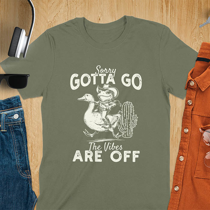A t-shirt with a frog in cowboy attire riding a goose. Text: "Sorry GOTTA GO" above and "The Vibes ARE OFF" below. This humorous tee-shirt features a funny frog tee and silly funny goose, a weird graphics shirt. A sarcastic meme shirt and funny genz meme, with rodeo cowboy and cowgirl t shirt elements. A vintage graphic tee and western style, an animal person gift. A funny unhinge shirt or mom gift alternative.