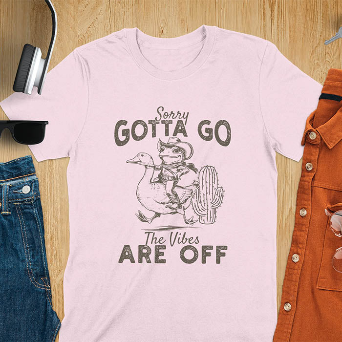 A t-shirt with a frog in cowboy attire riding a goose. Text: "Sorry GOTTA GO" above and "The Vibes ARE OFF" below. This humorous tee-shirt features a funny frog tee and silly funny goose, a weird graphics shirt. A sarcastic meme shirt and funny genz meme, with rodeo cowboy and cowgirl t shirt elements. A vintage graphic tee and western style, an animal person gift. A funny unhinge shirt or mom gift alternative.