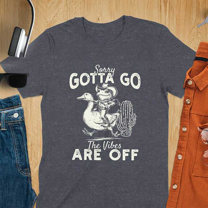 A t-shirt with a frog in cowboy attire riding a goose. Text: "Sorry GOTTA GO" above and "The Vibes ARE OFF" below. This humorous tee-shirt features a funny frog tee and silly funny goose, a weird graphics shirt. A sarcastic meme shirt and funny genz meme, with rodeo cowboy and cowgirl t shirt elements. A vintage graphic tee and western style, an animal person gift. A funny unhinge shirt or mom gift alternative.