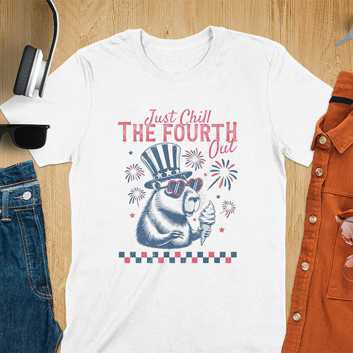 A t-shirt with the text 'Just Chill The Fourth Out' in red and blue lettering. A cartoon illustration of a capybara wearing a red, white, and blue top hat and sunglasses and holding an ice cream cone is below the text. The background of the illustration is a red, white, and blue fireworks display.