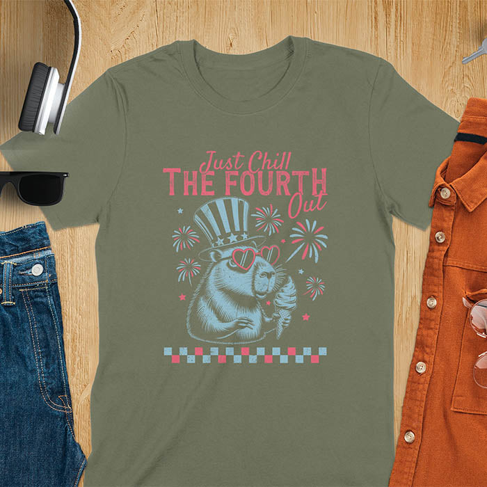 A t-shirt with the text 'Just Chill The Fourth Out' in red and blue lettering. A cartoon illustration of a capybara wearing a red, white, and blue top hat and sunglasses and holding an ice cream cone is below the text. The background of the illustration is a red, white, and blue fireworks display.