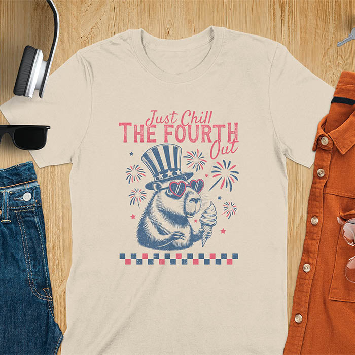 A t-shirt with the text 'Just Chill The Fourth Out' in red and blue lettering. A cartoon illustration of a capybara wearing a red, white, and blue top hat and sunglasses and holding an ice cream cone is below the text. The background of the illustration is a red, white, and blue fireworks display.