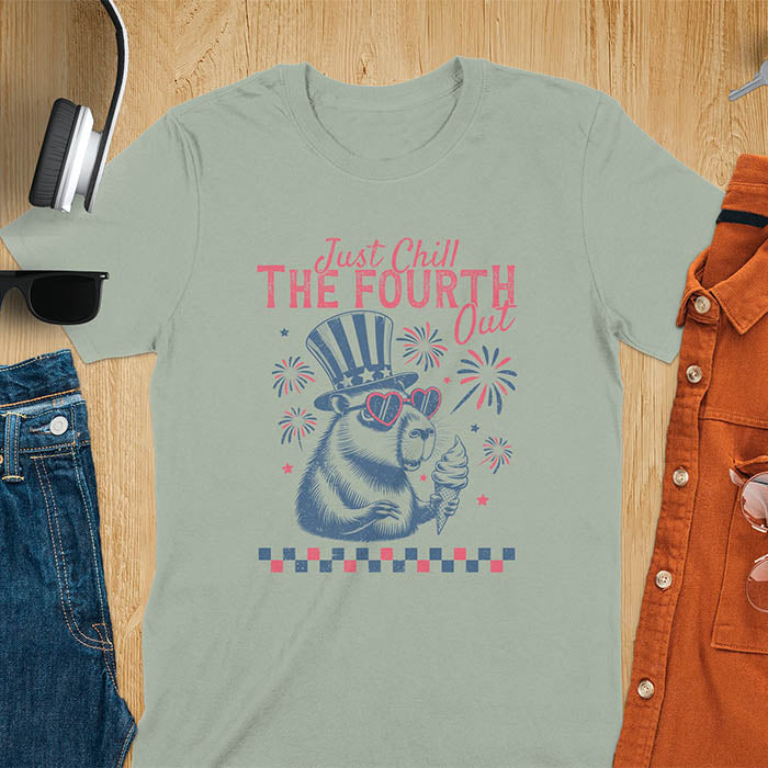 A t-shirt with the text 'Just Chill The Fourth Out' in red and blue lettering. A cartoon illustration of a capybara wearing a red, white, and blue top hat and sunglasses and holding an ice cream cone is below the text. The background of the illustration is a red, white, and blue fireworks display.