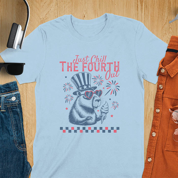 A t-shirt with the text 'Just Chill The Fourth Out' in red and blue lettering. A cartoon illustration of a capybara wearing a red, white, and blue top hat and sunglasses and holding an ice cream cone is below the text. The background of the illustration is a red, white, and blue fireworks display.