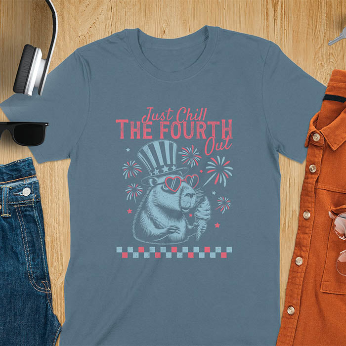 A t-shirt with the text 'Just Chill The Fourth Out' in red and blue lettering. A cartoon illustration of a capybara wearing a red, white, and blue top hat and sunglasses and holding an ice cream cone is below the text. The background of the illustration is a red, white, and blue fireworks display.
