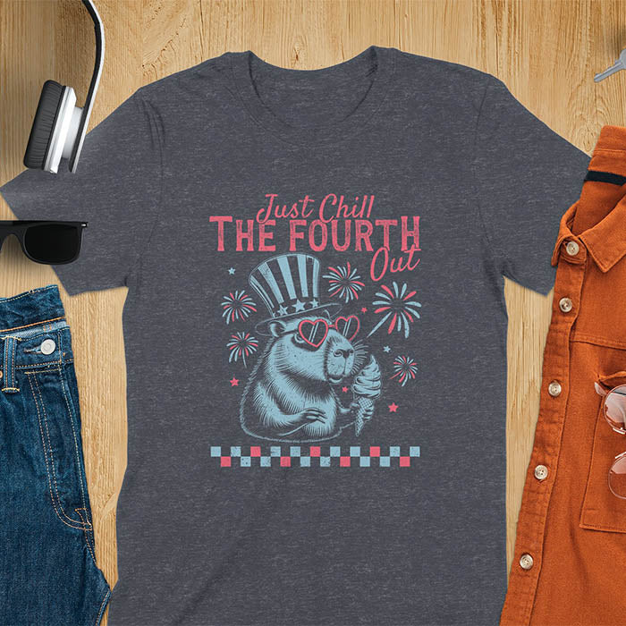 A t-shirt with the text 'Just Chill The Fourth Out' in red and blue lettering. A cartoon illustration of a capybara wearing a red, white, and blue top hat and sunglasses and holding an ice cream cone is below the text. The background of the illustration is a red, white, and blue fireworks display.