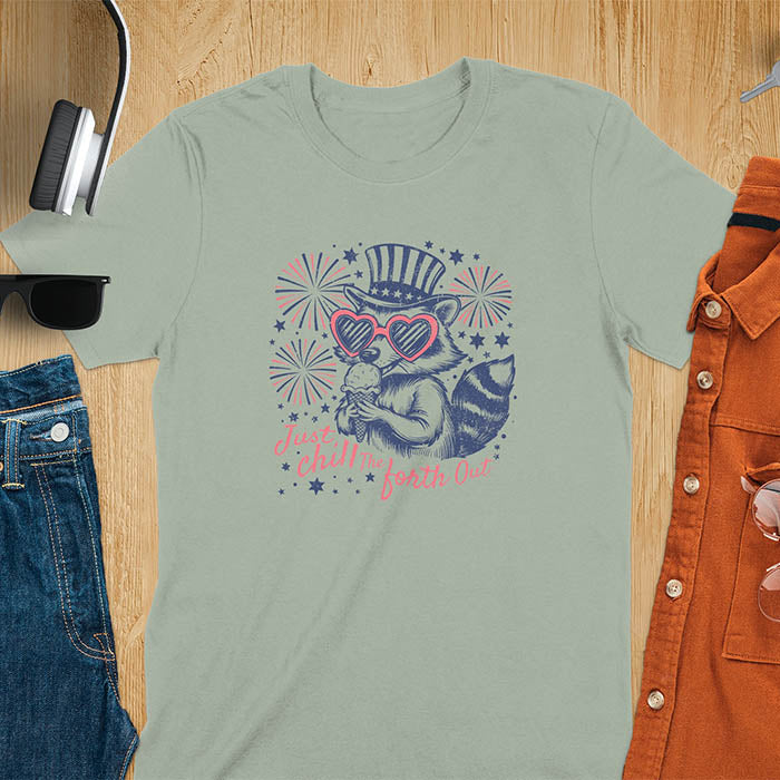 A t-shirt with a raccoon in a patriotic top hat & heart sunglasses, holding an ice cream cone. Fireworks burst around it. Text: "JUST CHILL THE FOURTH OUT". This raccoon cuteness tee is a funny 4th of July fourth of july shirt for chill the 4th out. A fireworks shirt, patriotic merica tee & freedom t shirt for independence day. Has vintage aesthetics, a funny patriot animal person gift or mom gift among 4th of july shirts.
