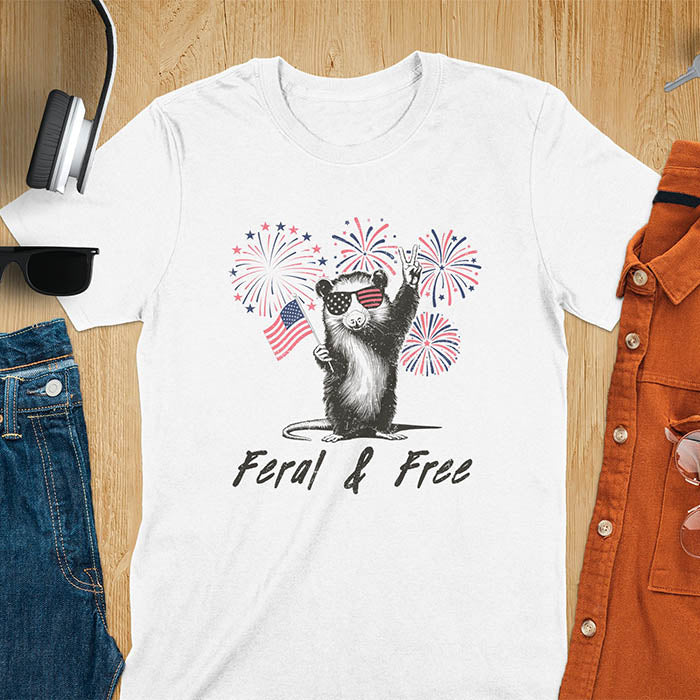 A t-shirt with an opossum wearing patriotic sunglasses. It holds a flag & makes a peace sign, with fireworks behind. Text: "Feral & Free". This unhinged meme tshirt is a merica shirt for Funny 4th of July. A summer tee-shirt with American pride shirt & feral tee-shirt vibes. Features opossum & animal cuteness tee, a feral raccoon shirt for USA Independence Day. A retro america t shirt as an animal person gift or gift for mom.