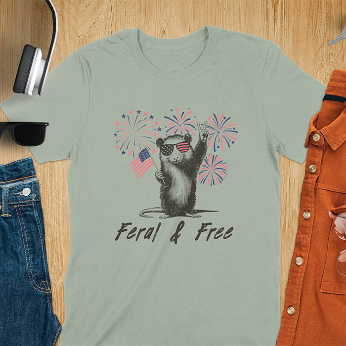 A t-shirt with an opossum wearing patriotic sunglasses. It holds a flag & makes a peace sign, with fireworks behind. Text: "Feral & Free". This unhinged meme tshirt is a merica shirt for Funny 4th of July. A summer tee-shirt with American pride shirt & feral tee-shirt vibes. Features opossum & animal cuteness tee, a feral raccoon shirt for USA Independence Day. A retro america t shirt as an animal person gift or gift for mom.