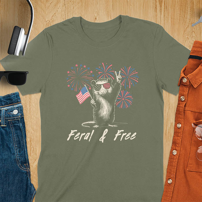 A t-shirt with an opossum wearing patriotic sunglasses. It holds a flag & makes a peace sign, with fireworks behind. Text: "Feral & Free". This unhinged meme tshirt is a merica shirt for Funny 4th of July. A summer tee-shirt with American pride shirt & feral tee-shirt vibes. Features opossum & animal cuteness tee, a feral raccoon shirt for USA Independence Day. A retro america t shirt as an animal person gift or gift for mom.