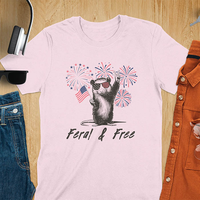 A t-shirt with an opossum wearing patriotic sunglasses. It holds a flag & makes a peace sign, with fireworks behind. Text: "Feral & Free". This unhinged meme tshirt is a merica shirt for Funny 4th of July. A summer tee-shirt with American pride shirt & feral tee-shirt vibes. Features opossum & animal cuteness tee, a feral raccoon shirt for USA Independence Day. A retro america t shirt as an animal person gift or gift for mom.