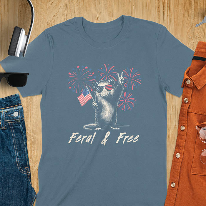 A t-shirt with an opossum wearing patriotic sunglasses. It holds a flag & makes a peace sign, with fireworks behind. Text: "Feral & Free". This unhinged meme tshirt is a merica shirt for Funny 4th of July. A summer tee-shirt with American pride shirt & feral tee-shirt vibes. Features opossum & animal cuteness tee, a feral raccoon shirt for USA Independence Day. A retro america t shirt as an animal person gift or gift for mom.