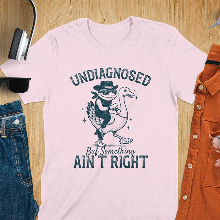 Funny cowboy frog riding goose t-shirt in light pink featuring cartoon frog in cowboy hat and boots riding a goose with text 'UNDIAGNOSED - But Something AIN'T RIGHT